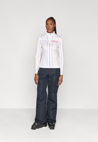 Long-sleeve, white zip-up top with vertical multi-colored stripes, paired with dark cargo pants and black ski boots. Simple design, functional fit.