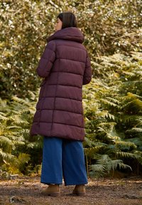 Bordeaux gekleed padded jas met hoge kraag, knielange ontwerp en brede mouwen. Gecombineerd met blauwe wijde jeans en bruine enkellaarsjes.