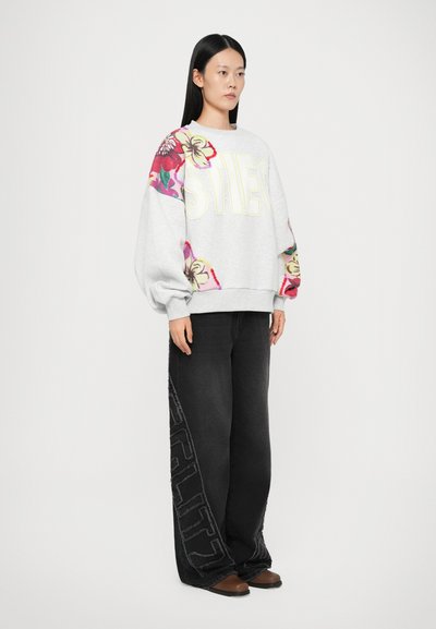 Gray sweatshirt with colorful floral patterns on sleeves and large "STLES" lettering. Black wide-leg jeans with contrasting text detail. Brown shoes.
