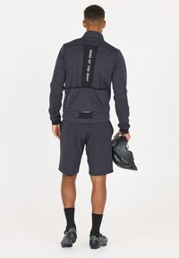 Männliches Radsportoutfit mit einer dunkelgrauen Jacke, die ein vertikales weißes Textdetail aufweist, kombiniert mit passenden Shorts und schwarzen Schuhen, während er einen Helm hält.