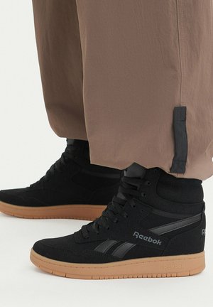 Sneakers Reebok nere alte con suola in gomma indossate con pantaloni marroni larghi su sfondo bianco.