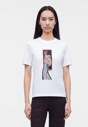 Femme aux longs cheveux noirs porte un t-shirt blanc avec une illustration abstraite colorée d’une silhouette, associé à un pantalon noir à motif chevron.