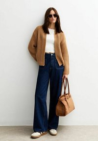 Cardigan di maglia marrone chiaro sopra una maglietta bianca, abbinato a jeans blu scuro a gamba larga e sneakers bianche. Borsa marrone con manici doppi.