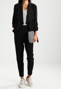 Schwarze, drapierte Jacke über einem grauen Oberteil, kombiniert mit schwarzen, taillierten Hosen und weißen Sneakers. Hält eine schwarz-weiße Clutch mit Zickzackmuster.