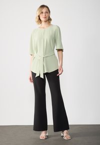 Top verde chiaro a maniche corte con un nodo in vita, abbinato a pantaloni neri a gamba larga e sandali bianchi con tacco a blocco. Design semplice.