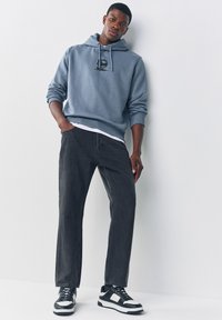 Niebieska bluza z logo, zestawiona z czarnymi jeansami i czarno-białymi trampkami. Bluza ma kaptur i ściągacze na rękawach, luźny krój.