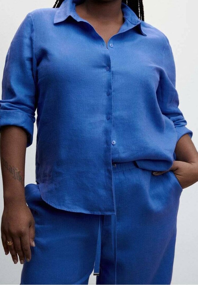 Chemise en lin bleue à boutons, manches retroussées, offrant une coupe décontractée. Le tissu a une texture lisse, assortie à un pantalon ample et assorti.