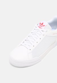 Bílé sportovní tenisky s síťovaným svrškem, hladkými syntetickými akcenty, růžovým logem, strukturovanou podrážkou a tradičním šněrováním.