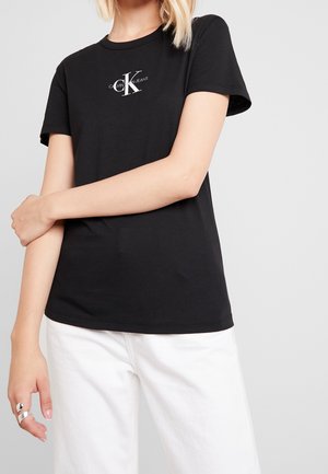 Kvinde iført en sort Calvin Klein Jeans t-shirt og hvide bukser, stående med armene over kors foran en ensfarvet baggrund.