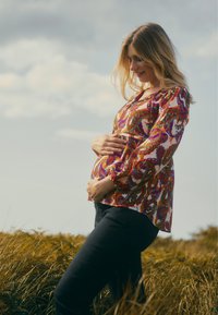 Kleurrijke blouse met paisleymotiv en lange mouwen, gecombineerd met donkere broek. De stof lijkt lichtgewicht en is ontworpen voor een zwangere silhout. 