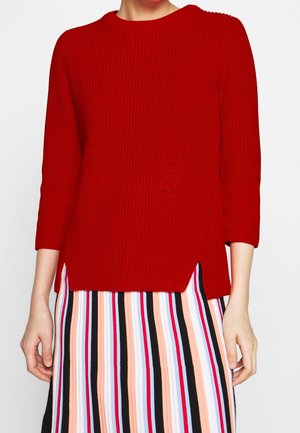 Personne portant un pull en tricot rouge à manches trois-quarts et une jupe à rayures verticales multicolores avec des rayures noires, blanches, roses, rouges et bleues.