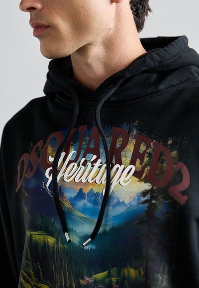 Giovane con un hoodie nero che presenta un paesaggio montano e la scritta "DSQUARED2 Heritage" stampata sul davanti.