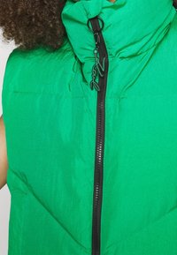 Groen puffer-vest met een hoge kraag, voorzien van een zwarte rits. De stof heeft een gladde, lichte textuur en bevat een ontwerperslabel aan de rits.