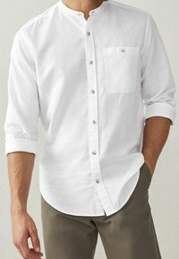 Bílá lněná košile s mandarínským límcem, dlouhými rukávy vyhrnutými, sedmi knoflíky vpředu a jednou náprsní kapsou se zapínáním na knoflík.