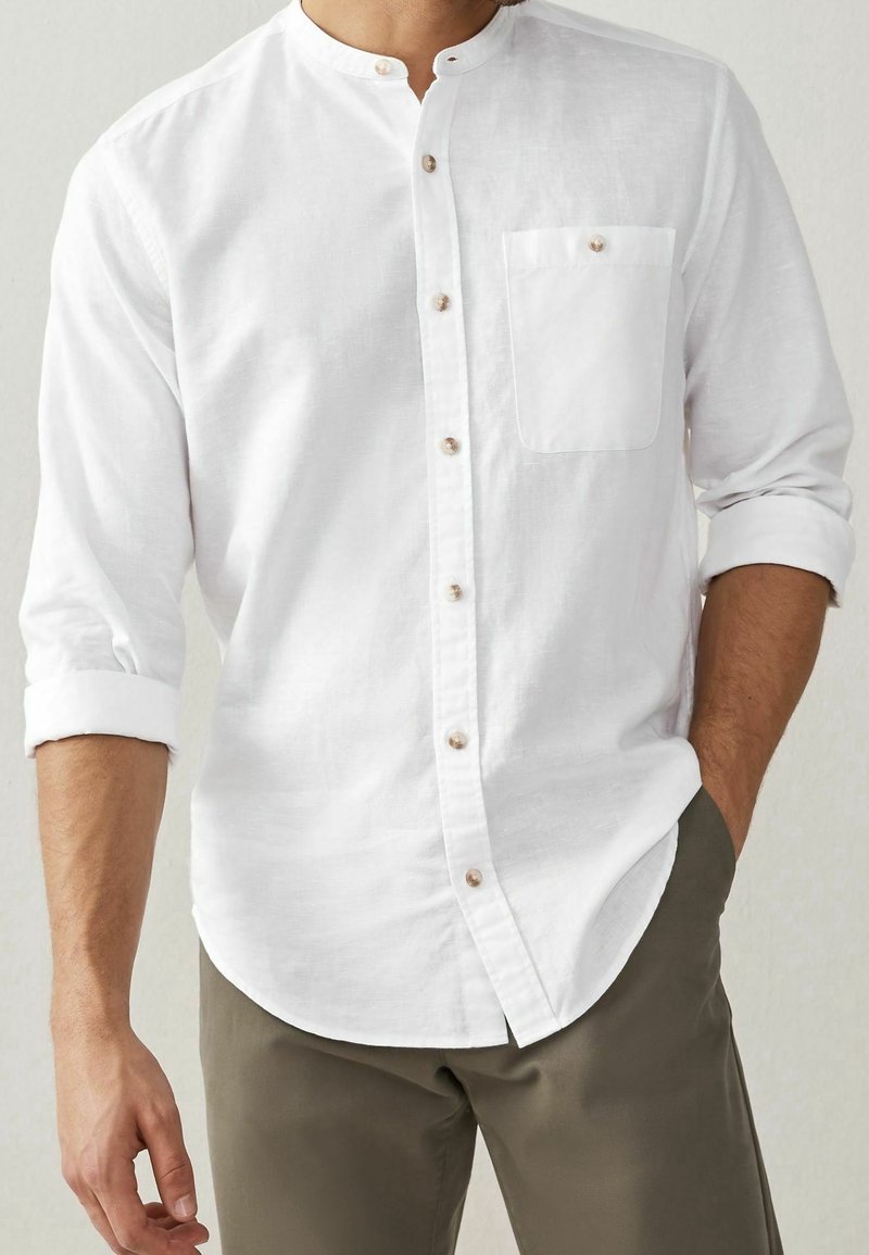 Bílá lněná košile s mandarínským límcem, dlouhými rukávy vyhrnutými, sedmi knoflíky vpředu a jednou náprsní kapsou se zapínáním na knoflík.