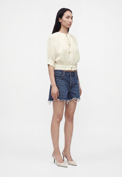 Femme portant un chemisier crème avec un nœud à l'avant et des manches bouffantes, un short en jean avec des bords effilochés, et des chaussures à talons beiges, debout de profil.