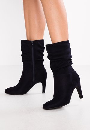Bottes en daim noir mi-mollet avec une coupe décontractée, fermeture éclair latérale, bout pointu et talons hauts fins portées sur des jambes nues sur fond blanc.