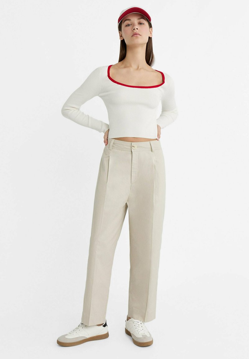 Maglietta bianca a costine con maniche lunghe e colletto rosso, abbinata a pantaloni beige plissettati e sneakers bianche con suole color testa di moro e dettagli neri.