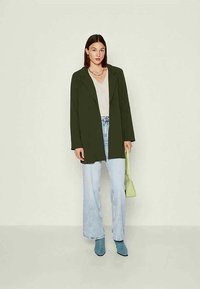 Γυναίκα που φοράει σκούρο πράσινο σακάκι, λευκή μπλούζα, ανοιχτό μπλε παντελόνι σε φαρδιά γραμμή, μπλε μποτάκια με τακούνι, κρατώντας ανοιχτό πράσινη τσάντα χεριού μπροστά σε απλό φόντο.