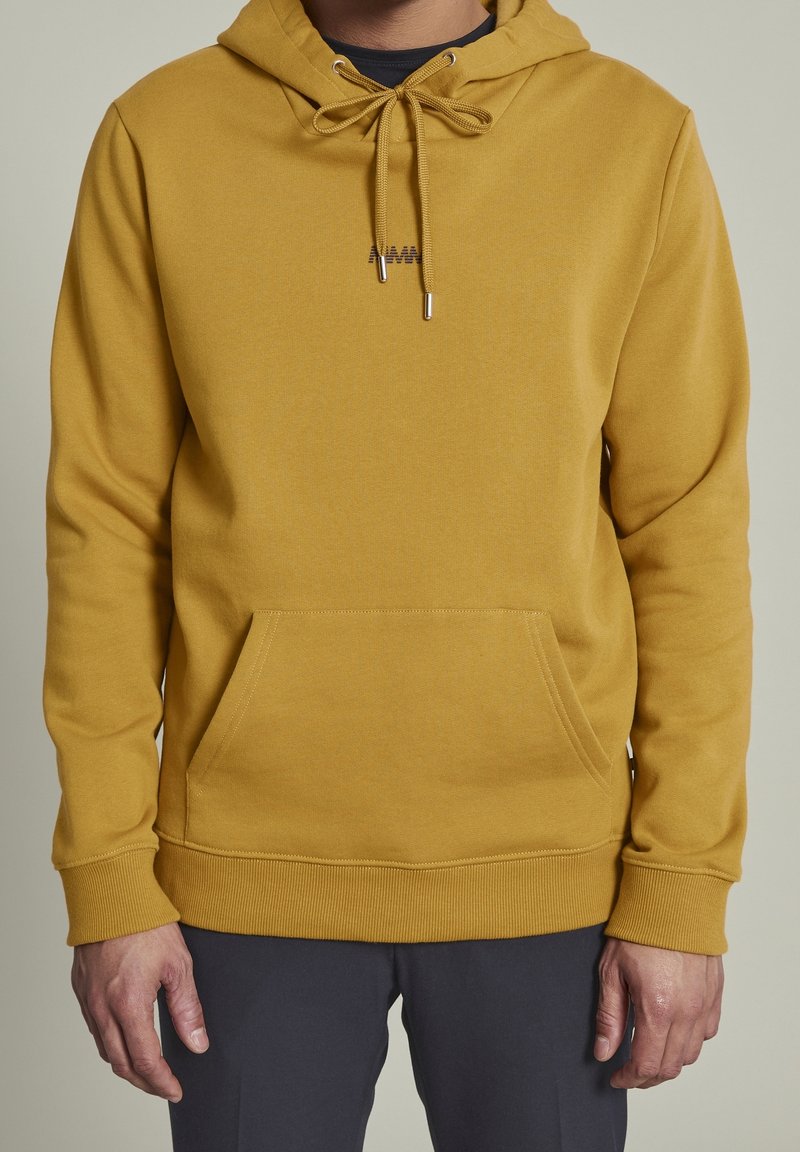 Homme portant un sweat à capuche jaune moutarde avec poche avant et cordons, associé à un pantalon foncé, visible des épaules aux hanches.