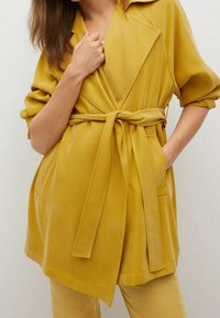 Manteau jaune ceinturé avec col, coupe oversize, manches retroussables et poches diagonales. Texture lisse avec une finition mate.