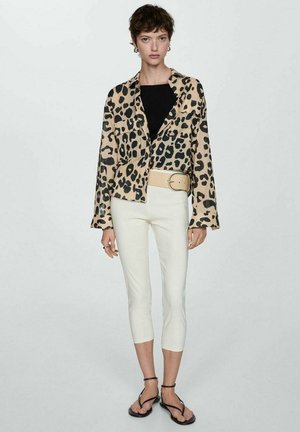 Femme portant une veste en imprimé léopard sur un haut noir, un pantalon blanc court avec une large ceinture beige, et des sandales noires à lanières, debout devant un fond uni.