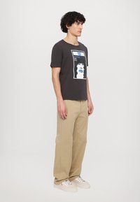 Donkergrijze t-shirt met een grafische print van een gezicht met blauwe lippen. Gecombineerd met beige wijde broek en witte sneakers.