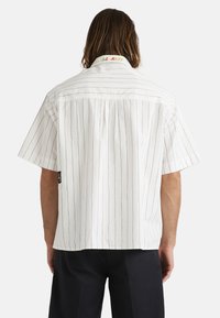 Witte blouse met korte mouwen en zwarte verticale strepen, met een kleurrijke geborduurde kraag en een klein label aan de linkerkant.