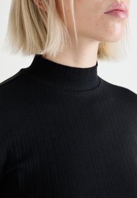 Svart ribbad turtleneck-top i elastisk tyg, med en figurnära design och hög krage. Slät yta med subtila vertikala linjer.