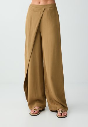 Vrouw draagt bruine, wijde broek met hoge taille en een overslagdetail aan de voorkant en beige sandalen tegen een effen lichte achtergrond.