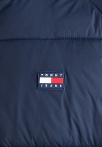 Tela de chaqueta acolchada de color azul marino, que presenta una etiqueta rectangular de Tommy Jeans con secciones blancas y rojas, con detalles cosidos a lo largo de las costuras.