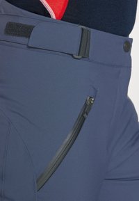 Mornarsko modre vodoodporne hlače z gladko teksturo, pasom na ježka in z zadrgo na strani, ki ima črno zadrgo.