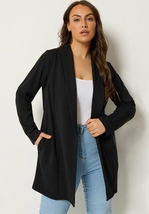 Vrouw draagt een zwarte blazer met open voorzijde, witte top en lichtblauwe spijkerbroek, staand tegen een lichte achtergrond met één hand in de broekzak.