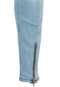 Hele sinine teksapüksid on kitsas lõikega, veidi kitsenevate säärtega ja välise pahkluu serval on nähtav tõmblukudetail.