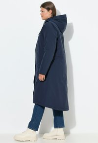 Námořnický dlouhý kabát s kapucí, vyrobený z hladké tkaniny. Disponuje kapsami na zip a má rovný siluetu, kombinovaný s béžovými kotníkovými botami.