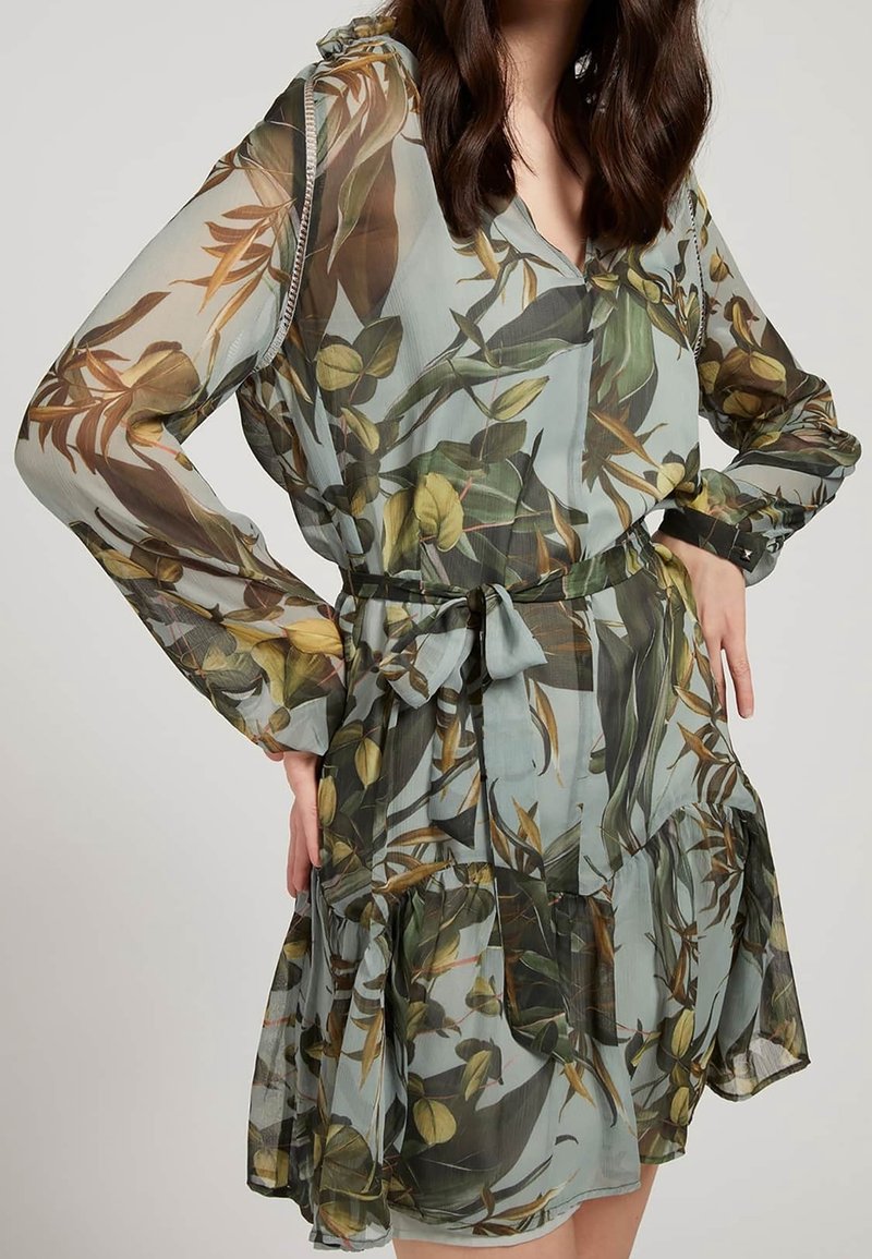Vestido floral hecho de una tela ligera y transparente, con un estampado botánico en verde y beige, mangas largas, escote en V y cintura ajustable con lazo.