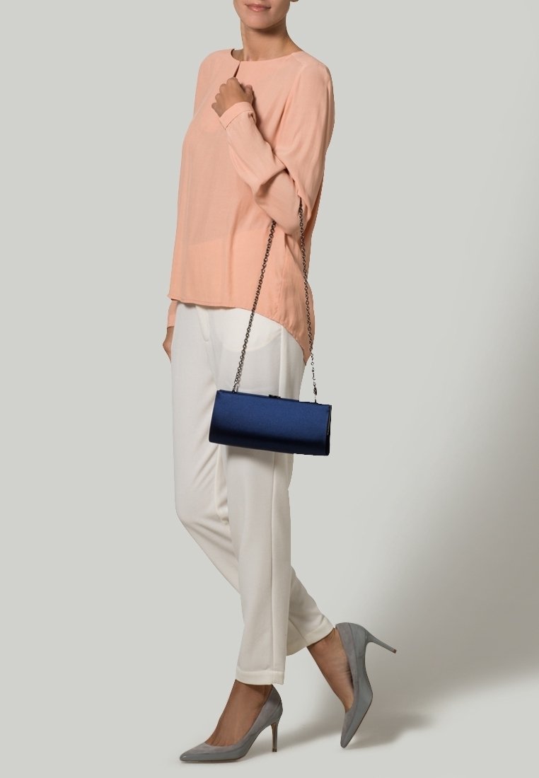 Kvinna i persikofärgad blus, vita byxor, grå högklackade skor, som håller en liten blå handväska med kedjesnor över axeln.