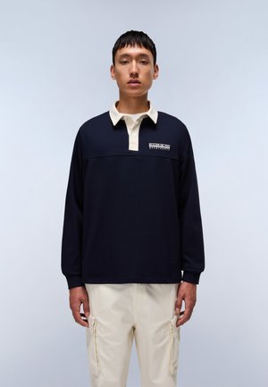 Marineblå langermet polo med kremfarget krage og mansjetter, med en logodesign på brystet. Båret med lysebeige cargo bukser.