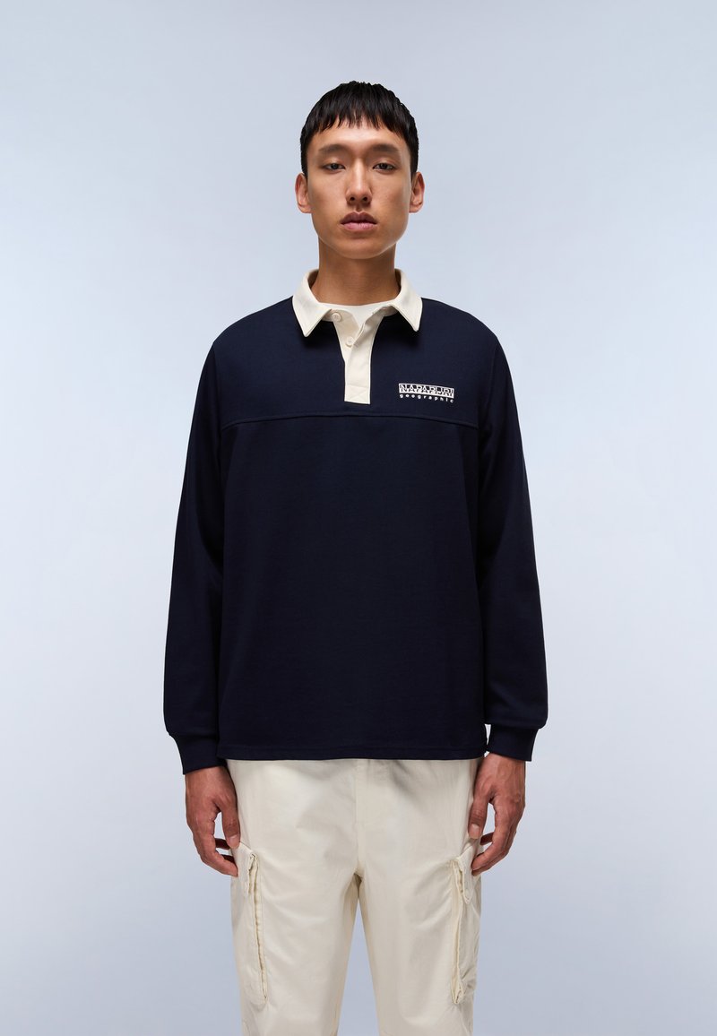 Polo de manga larga en color marino con cuello y puños de color crema, que presenta un diseño de logo en el pecho. Combinado con pantalones cargo de color beige claro.