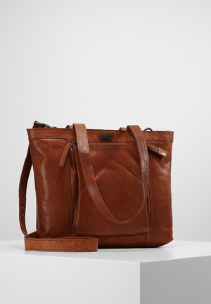 Sac cabas en cuir marron avec deux poignées, poche avant zippée et bandoulière amovible sur un piédestal blanc contre un fond uni.