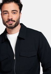 Veste noire à fermeture éclair sur le devant, en tissu texturé, avec deux poches sur la poitrine et un col classique. Portée par-dessus un vêtement blanc.