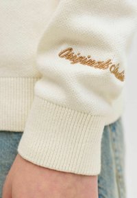 Poignet de pull en maille crème avec ourlet côtelé et texte brodé partiel en camel "Originals" au-dessus du poignet, porté avec un jean en denim bleu.