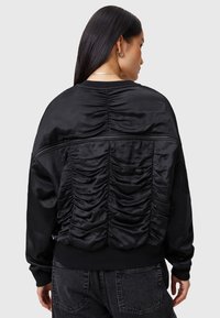 Giacca nera in satin con dettagli arricciati sulla schiena, polsini a costine e vestibilità rilassata. Texture liscia con una cucitura superiore con zip.