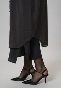 Femeie îmbrăcată cu ciorapi negri transparenți, pantofi cu toc înalt, vârful ascuțit, cu curele la gleznă, și o fustă asimetrică neagră.