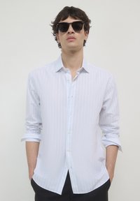 Chemise blanche à manches longues avec de subtiles rayures verticales, col boutonné et manches retroussées, assortie à des lunettes de soleil foncées.