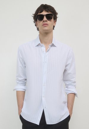 Camicia bianca a maniche lunghe con sottili righe verticali, colletto a bottone e maniche arrotolate, abbinata a occhiali da sole scuri.