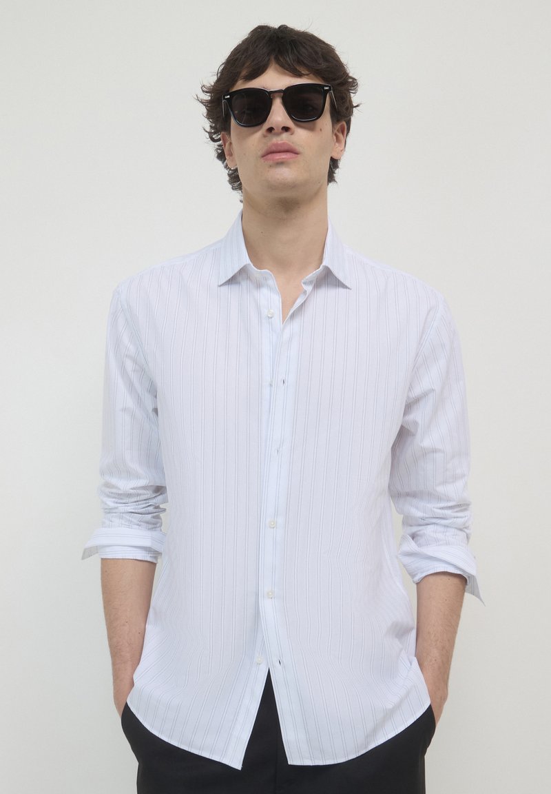 Chemise blanche à manches longues avec de subtiles rayures verticales, col boutonné et manches retroussées, assortie à des lunettes de soleil foncées.