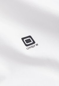 Witte stof met een zwart logo met een vierkante omtrek en het woord "ombre" in kleine letters. Soepele textuur, minimalistisch ontwerp.