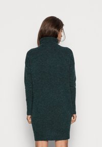 Abito maglione a maniche lunghe, di colore verde scuro, con colletto alto, polsini a costine e una vestibilità ampia, realizzato in un materiale morbido e testurizzato.