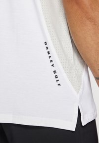 Chemise de golf blanche avec logo "OAKLEY GOLF" noir, dotée d'un panneau texturé gris. Les bords ourlés et les fentes latérales améliorent la respirabilité.